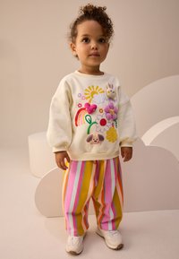 Sweatshirt crème avec appliqués colorés d'animaux et de fleurs, associée à un pantalon ample rayé rose, jaune et bleu. Baskets blanches.