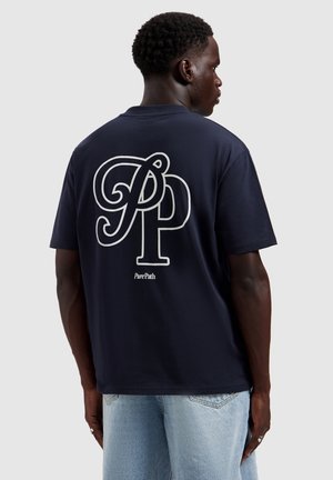 Man in een marineblauw T-shirt met grote verstrengelde "PP" letters en de tekst "Pure Path" op de achterkant, gecombineerd met lichtblauwe jeans.