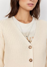 Béžový pletený cardigan s výstřihem do V a třemi dřevěnými knoflíky, nošený přes světlé tričko. Detailní záběr ukazuje texturu a detaily knoflíků.