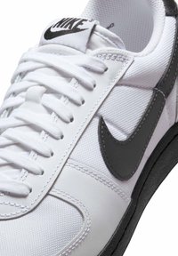 Nike tenisice s gornjim dijelom od bijelog tekstila, sivim kožnim swooshom, crnom gumenom potplatom i bijelim vezicama. Imaju podstavljeni jezik i teksturiranu mrežicu.