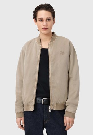 Young person with short brown hair wears a beige zip-up bomber jacket over a black shirt and dark jeans with belt, standing against plain background.