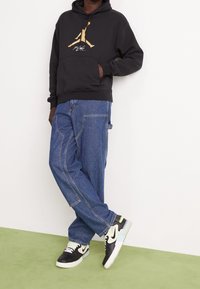 Persona che indossa un cappuccio nero con il logo Jumpman dorato, jeans blu larghi e scarpe Nike nere e bianche, in piedi contro un muro bianco e un pavimento verde.