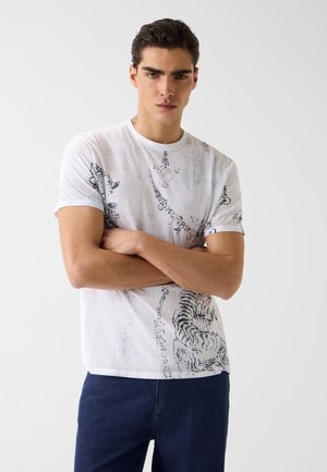 Giovane uomo con le braccia incrociate che indossa una maglietta bianca con disegni astratti di animali neri e scritte, abbinata a pantaloni blu scuro.