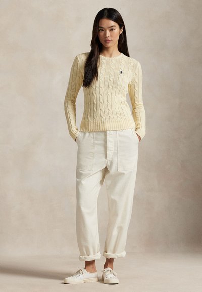 Donna in piedi che indossa un maglione color crema a treccia, pantaloni bianchi larghi con risvolti e scarpe da ginnastica bianche con lacci, su uno sfondo semplice.