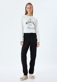 Camisa blanca de manga larga con texto negro que dice "El FIN DE SEMANA es para dormir." Pantalones negros con cintura elástica. Zapatillas de casa de piel beige. Diseño simple.