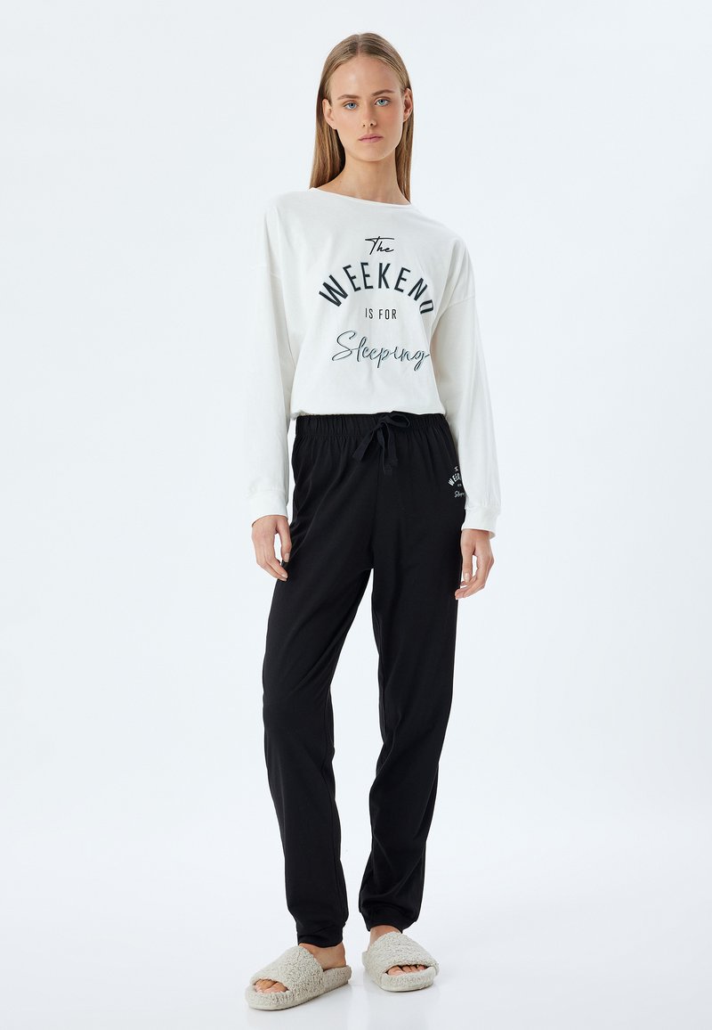 Camisa blanca de manga larga con texto negro que dice "El FIN DE SEMANA es para dormir." Pantalones negros con cintura elástica. Zapatillas de casa de piel beige. Diseño simple.