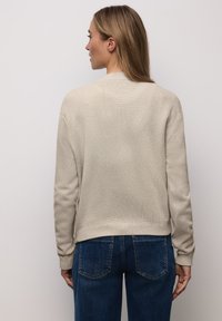 Hellbeige Strick-Cardigan mit strukturierter Stoffoberfläche, gerippten Bündchen und Saum. Draußen getragen mit dunkelblauen Jeans, die von hinten eine entspannte Silhouette zeigen.
