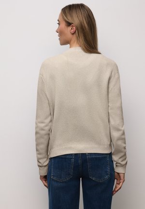 Hellbeige Strick-Cardigan mit strukturierter Stoffoberfläche, gerippten Bündchen und Saum. Draußen getragen mit dunkelblauen Jeans, die von hinten eine entspannte Silhouette zeigen.
