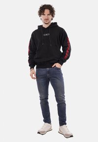 Felpa nera con grafica rossa sulle maniche, cappuccio e logo frontale. Indossata con jeans blu e sneakers beige, in piedi contro uno sfondo bianco.