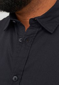 Camisa negra de cuello con una textura de tela suave, que presenta un cierre de botones y un sutil detalle de tapeta cerca del cuello.