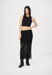Fustă din dantelă neagră cu un design transparent, asortată cu un top scurt negru. Modelul poartă cizme negre până la genunchi. Fundal minimalist.