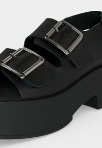 Μαύρο πέδιλο με πλατφόρμα με δύο φαρδιές δερμάτινες λουρίδες και ασημένιες ορθογώνιες μεταλλικές αγκράφες.