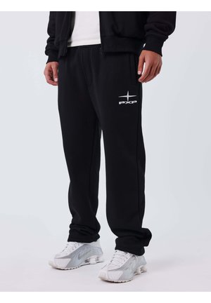 Homme portant un pantalon de jogging noir avec le logo blanc PXP sur la jambe gauche et des chaussures de sport blanches, debout devant un fond uni.