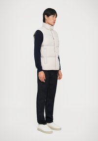 Hellbeige gesteppte Weste mit hohem Kragen, Knopfverschluss vorne und Seitentaschen, getragen über einem dunklen Pullover und kombiniert mit dunklen Jeans und weißen Sneakers.