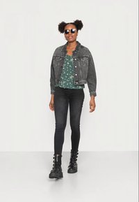 Jaqueta de ganga preta sobre uma blusa floral verde, combinada com jeans skinny pretos e botas de tornozelo pretas com atacadores e detalhes metálicos.