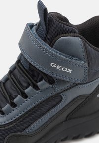 Zapato azul oscuro con parte superior de malla texturizada y acentos de cuero suave. Presenta una correa de velcro, costuras en contraste y una suela negra gruesa.