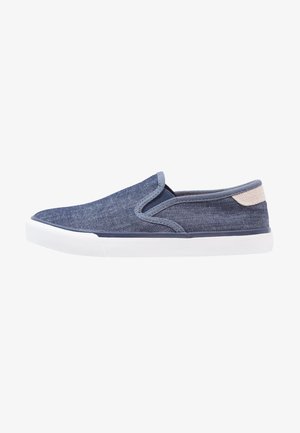Vapaa-ajan slip-on-tennari sinisestä farkkukankaasta, valkoisella kumipohjalla ja beigenvärisellä kantapäälenkillä, sivuprofiilinäkymä.
