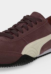 Baskets en daim marron foncé avec des lacets marron épais, une bande blanche incurvée sur le côté et une semelle extérieure en caoutchouc noir texturé.