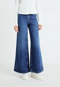Wijd gesneden denim jeans in donkerblauw, met een rechte snit en franjes aan de onderkant. Ggedragen met een witte ribbeltop en witte sneakers.