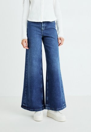 Wijd gesneden denim jeans in donkerblauw, met een rechte snit en franjes aan de onderkant. Ggedragen met een witte ribbeltop en witte sneakers.