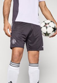 Čierne športové šortky s emblémom Manchester United, kombinované s bielymi podkolienkami s čiernymi pruhmi a vzorovanou futbalovou loptou.