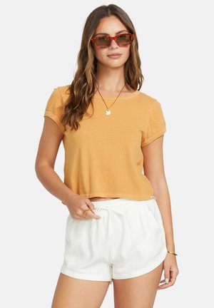 Frau trägt ein oranges, kurz geschnittenes T-Shirt, weiße Shorts mit Kordelzug, Sonnenbrille und schlichten Goldschmuck, steht vor einem einfarbigen Hintergrund.