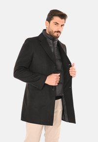 Uomo con pantaloni beige e cappotti neri stratificati, che tiene aperto il cappotto esterno, su sfondo grigio.