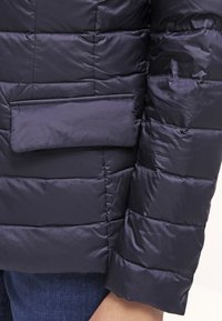 Närbild på en person som bär en svart vadderad pufferjacka med en horisontell ficka och ärm synlig över blå jeans.