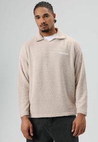 Beige fleece pullover met een kraag, lange mouwen en een getextureerd oppervlak. Heeft een klein wit logo op de borst.