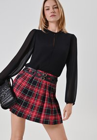 Schwarze, durchsichtige Bluse mit langen Ärmeln, kombiniert mit einem roten und schwarzen Karomuster-Rock mit Falten, der einen schwarzen Gürtel mit doppelten Schnallen hat.
