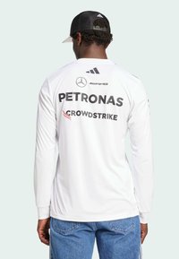 Camiseta deportiva blanca de manga larga con estampados de logotipos en negro: "PETRONAS" y "CROWDSTRIKE", con cuello redondo y puños acanalados.