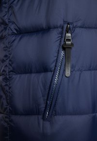 Granatowa kurtka puchowa z poziomym przeszyciem, strukturalnym materiałem i kieszenią zapinaną na zamek. Zamek ma uchwyt z materiału.