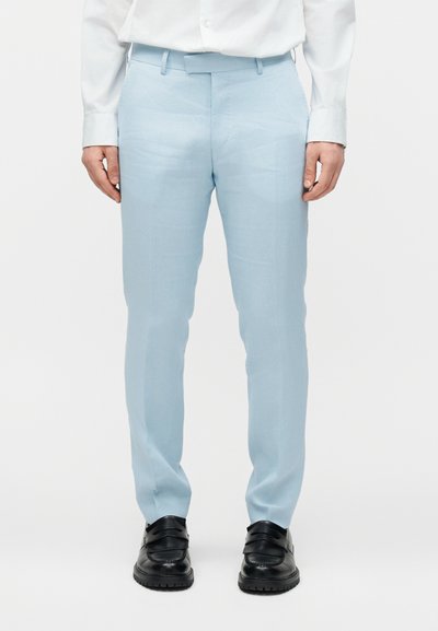 Άνδρας με ανοιχτό μπλε παντελόνι, λευκό πουκάμισο και μαύρα δερμάτινα μοκασίνια, στέκεται μπροστά σε λευκό φόντο.