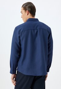 Camisa azul marino de manga larga hecha de tejido texturizado, con cuello puntiagudo, puños abotonados y un corte relajado. El patrón es sutil.