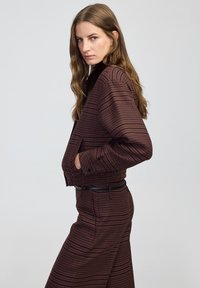 Giacca marrone a fantasia con un design corto, dotata di un colletto alto, tasche frontali e una cintura aderente. Abbinata a pantaloni coordinati.