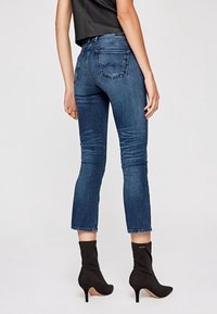 Tmavě modré zkrácené džíny vyrobené z denimu, s těsným střihem, zadními kapsami a viditelným šitím. Nosené s černými vysokými podpatky.