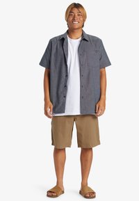 Camisa de botón de manga corta, gris oscuro, camiseta blanca debajo, pantalones cortos color beige y sandalias marrones. La camisa tiene un bolsillo en el pecho y un corte clásico.