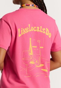 Vaaleanpunainen puuvillapaita, jossa on keltainen grafiikka viinilasista ja pulloista takana, ja tekstinä "LikeLocalsDo" designin yläpuolella.