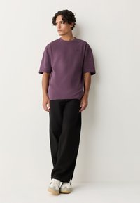 Lila T-Shirt mit kurzen Ärmeln und rundem Ausschnitt, geripptes Material, kombiniert mit schwarzen Hosen mit weitem Bein und grau-weißen Sneakern mit heller Sohle.