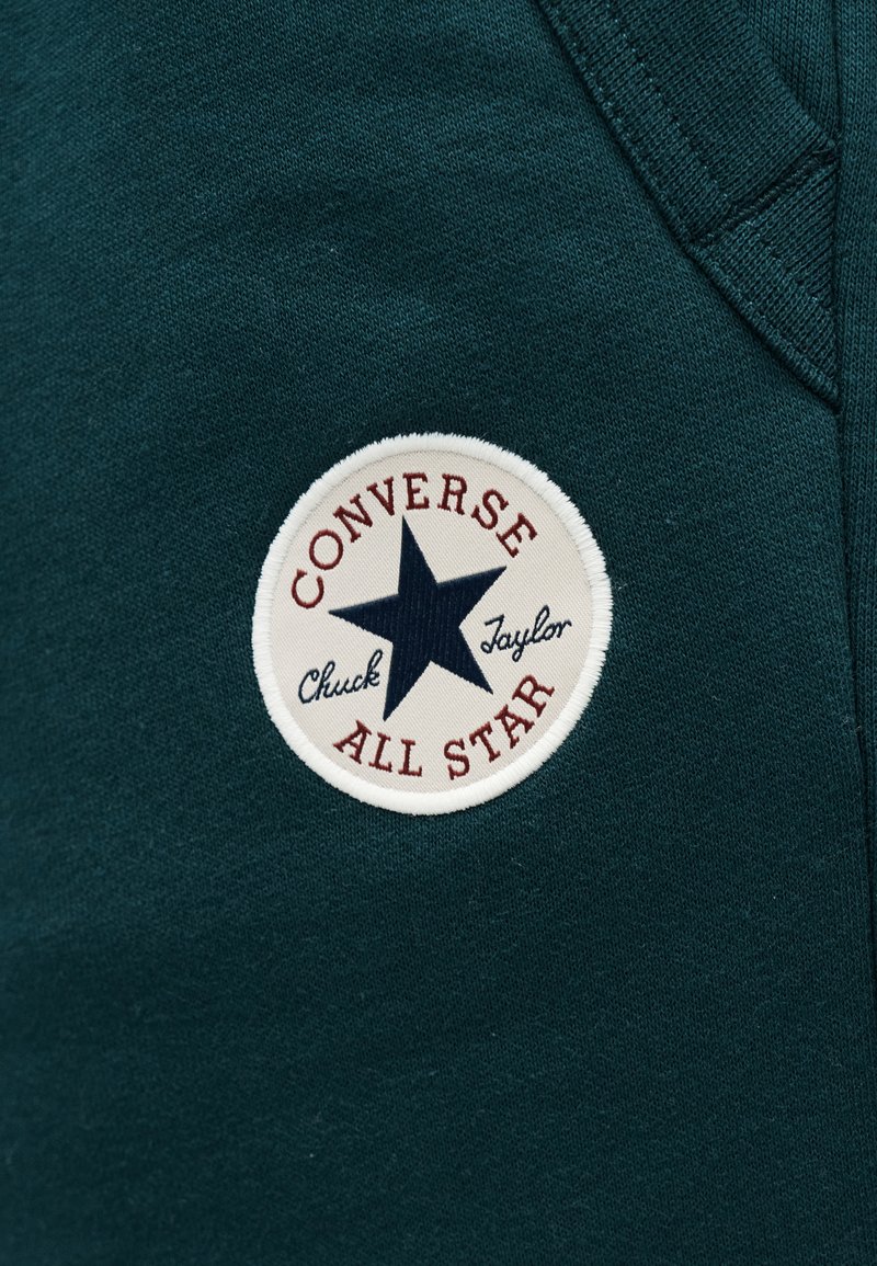Dark green fabric with a round white patch featuring a black star and text "Converse Chuck Taylor All Star" embroidered.