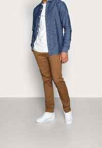 Codzienny strój składający się z ciemnoniebieskiej koszuli zapinanej na guziki, białego t-shirtu, brązowych spodni oraz białych sneakersów, podkreślający kontrast tkanin i kolorów.