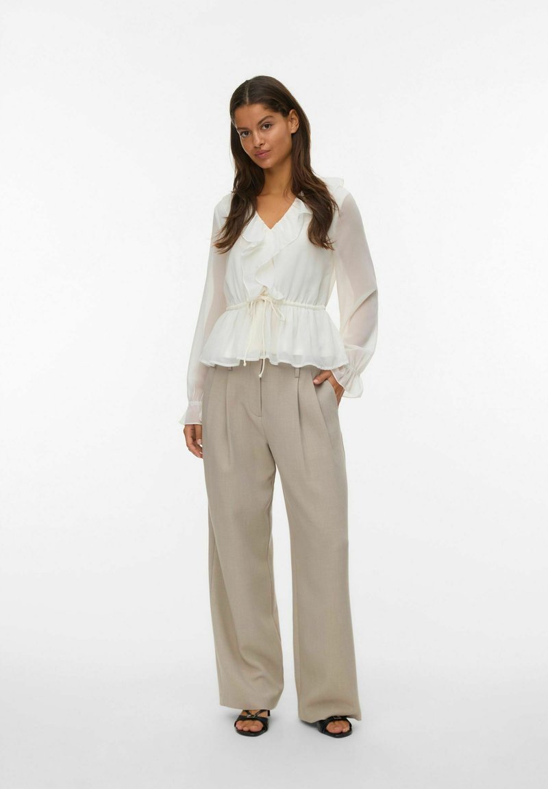 Hvit semi-gjennomsiktig bluse med volangkrage, snor i midjen og lange ermer, kombinert med lyse beige vide bukser. Sorte sandaler.