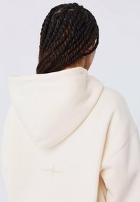 Persoon met lang, verstrengeld donker haar die een crèmekleurige hoodie draagt met een klein stersymbool op de achterkant, bekeken van achteren.