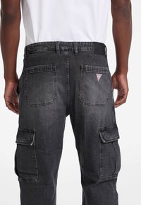 Pantaloni cargo din denim negru, cu buzunare laterale și pe spate, un logo aplicat pe spate și o croială lejeră. Caracteristici ale unei texturi de țesătură rezistente.