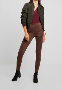 Veste bomber olive, haut bordeaux côtelé avec bordure en dentelle, leggings marron aspect daim et bottines noires à talons. Modèle en position debout.