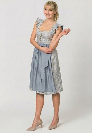 Blonde Frau lächelt, trägt ein blaues Blumenmusterkleid mit Rüschenärmeln und Schürze, steht mit einem Arm verschränkt und dem anderen erhoben.