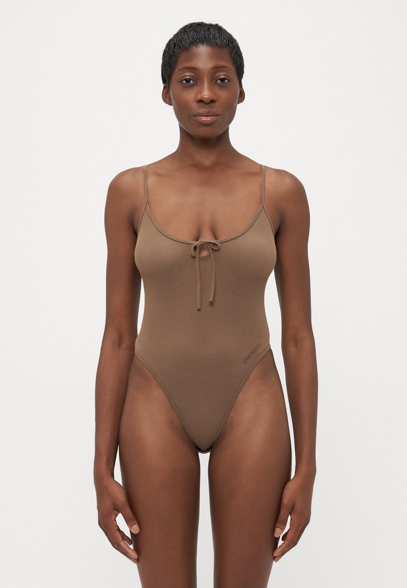 Femme aux cheveux courts portant un bodysuit côtelé brun avec des fines bretelles et un petit nœud à l'avant, se tenant devant un fond blanc uni.