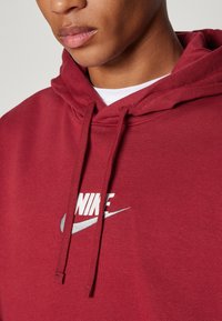 Sudadera con capucha roja que incluye un bolsillo frontal, capucha con cordón y un logo blanco de Nike con un acento plateado en forma de swoosh. Tela suave con acabado texturizado.