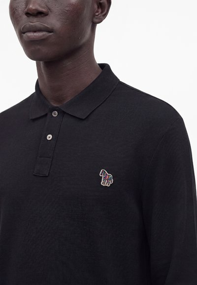 Gros plan d'une personne portant un polo noir avec un petit logo zèbre brodé en couleurs sur le côté gauche de la poitrine.