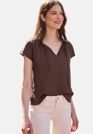 Kvinde smilende, iført en brun bluse med korte ærmer og V-hals samt cremefarvede bukser, stående mod en ensfarvet lys baggrund.